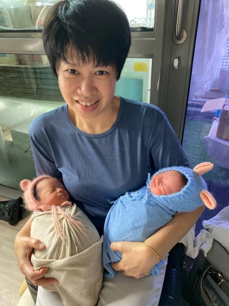 Photo: May Leng smiling, holding two swaddled newborn twins